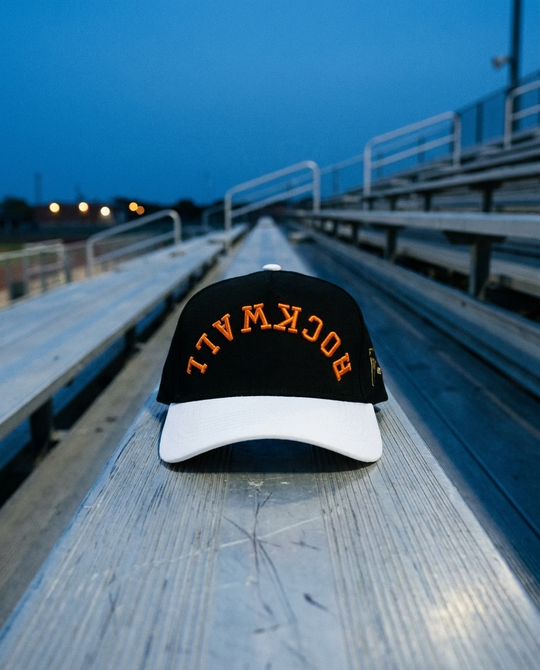 BKWHOR Rockwall upside down hat, black crown, white brim, orange embroidery, on stadium bleachers at blue hour in Rockwall Texas