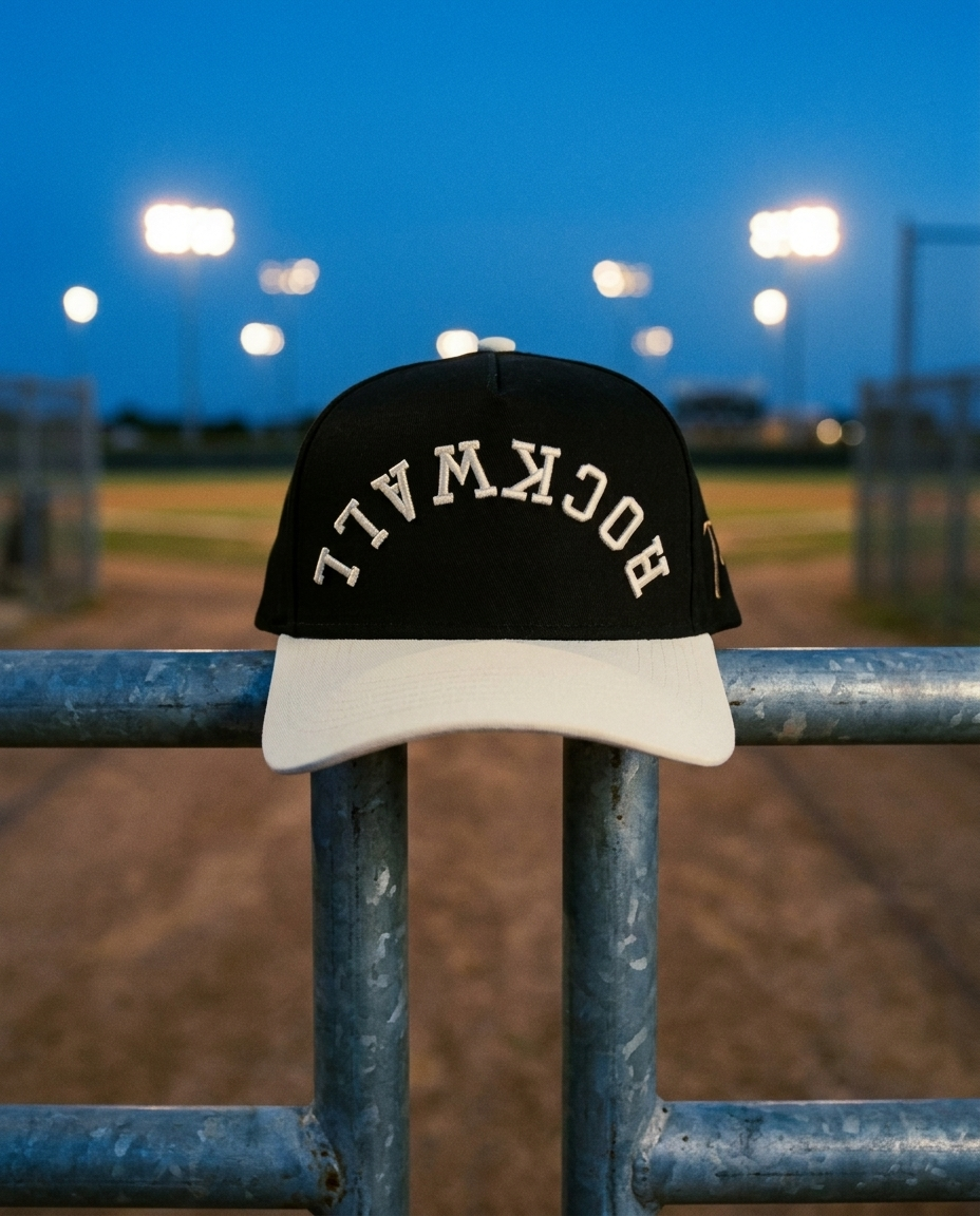BKCRCR Rockwall upside down hat, black crown, cream brim, cream embroidery, on a ball field gate rail at dusk in Rockwall Texas