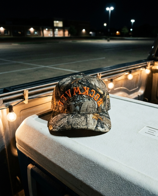 CMCMOR Rockwall upside down hat, camo crown and brim, orange embroidery, on a tailgate cooler at night in Rockwall Texas