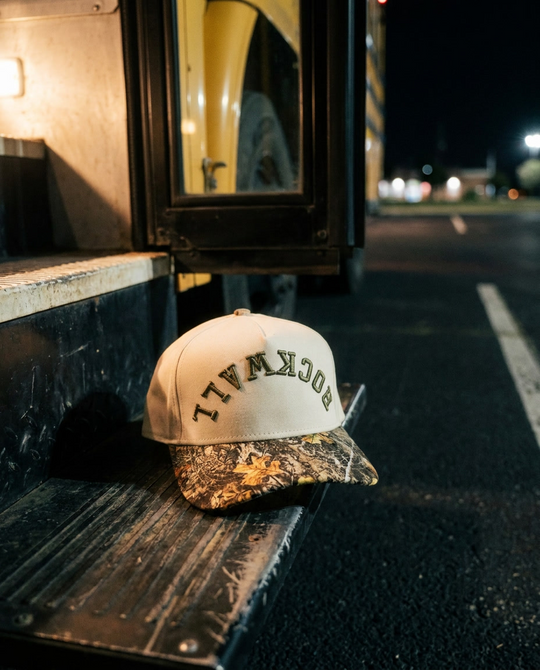 CRCMAG Rockwall upside down hat, cream crown, camo brim, army green embroidery, on a bus step at night in Rockwall Texas