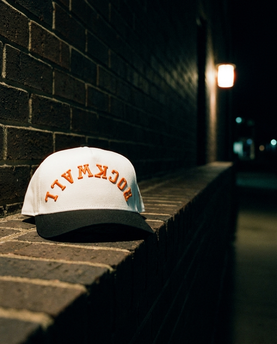 WHBKOR Rockwall upside down hat, white crown, black brim, orange embroidery, on a brick ledge at night in Rockwall Texas
